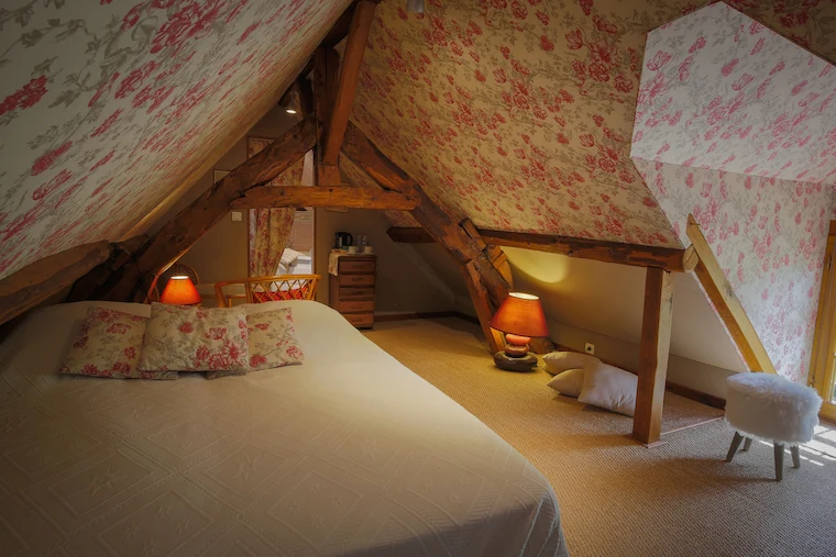 Chambre mansardée sous combles avec poutres apparentes et papier peint fleuri, lit double et coin salon cosy