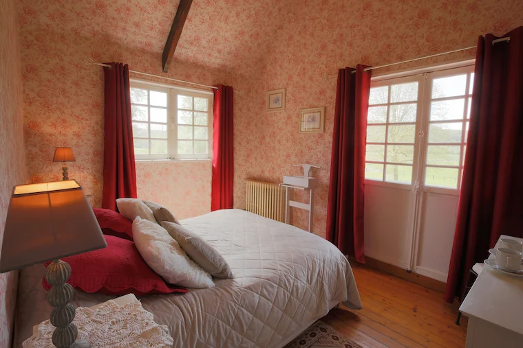 Chambre romantique au papier peint fleuri rose et rideaux rouges, avec lit double et fenêtres sur la campagne